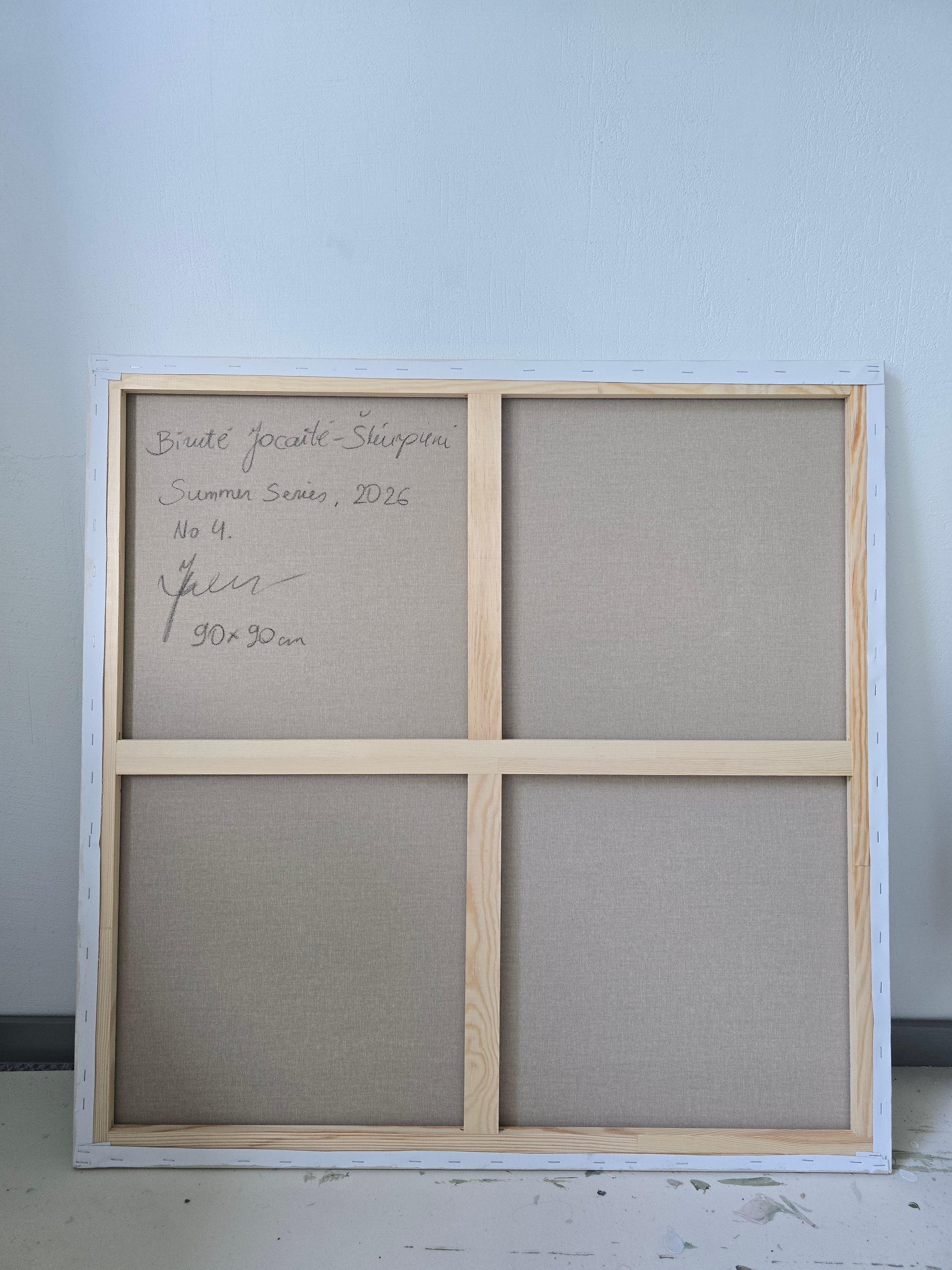 A square four-panel wooden canvas or wall art frame with beige fabric panels, photographed from above against a light gray wall. Handwritten text appears in the lower left panel.