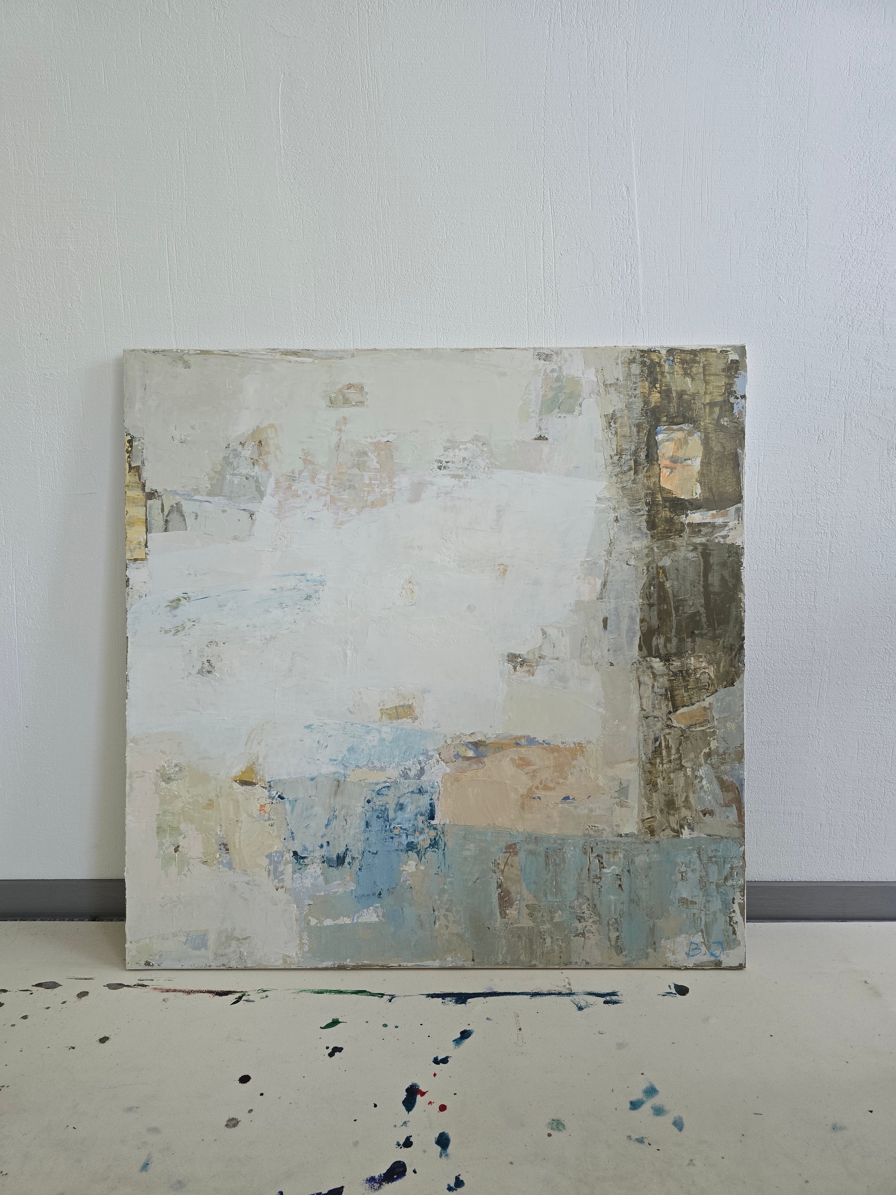 An abstract square canvas painting with layered textures in off-white, neutral, beige, light blue, and brown displayed against a white wall.