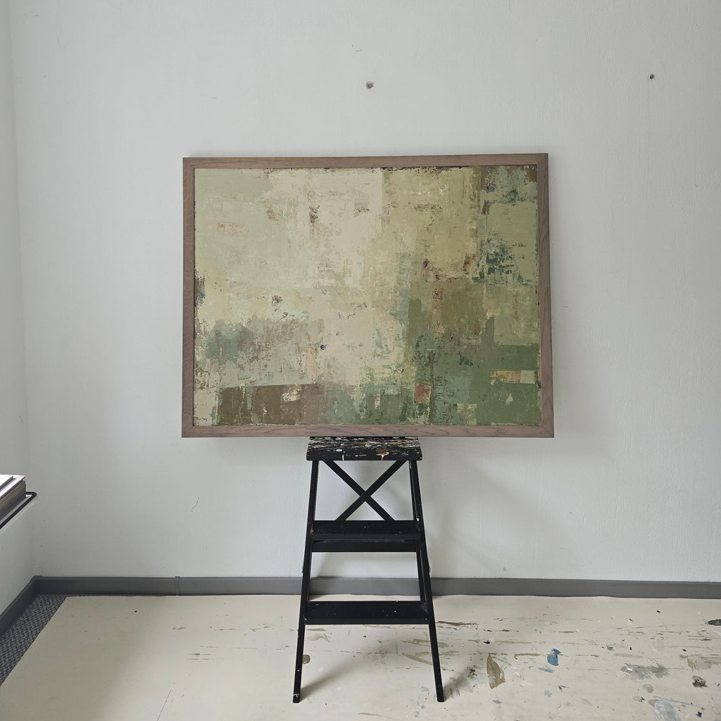 A square framed art original with a textured, distressed patchwork design in muted green, beige, and brown tones, shown hanging on a plain white wall next to a black metal stool.