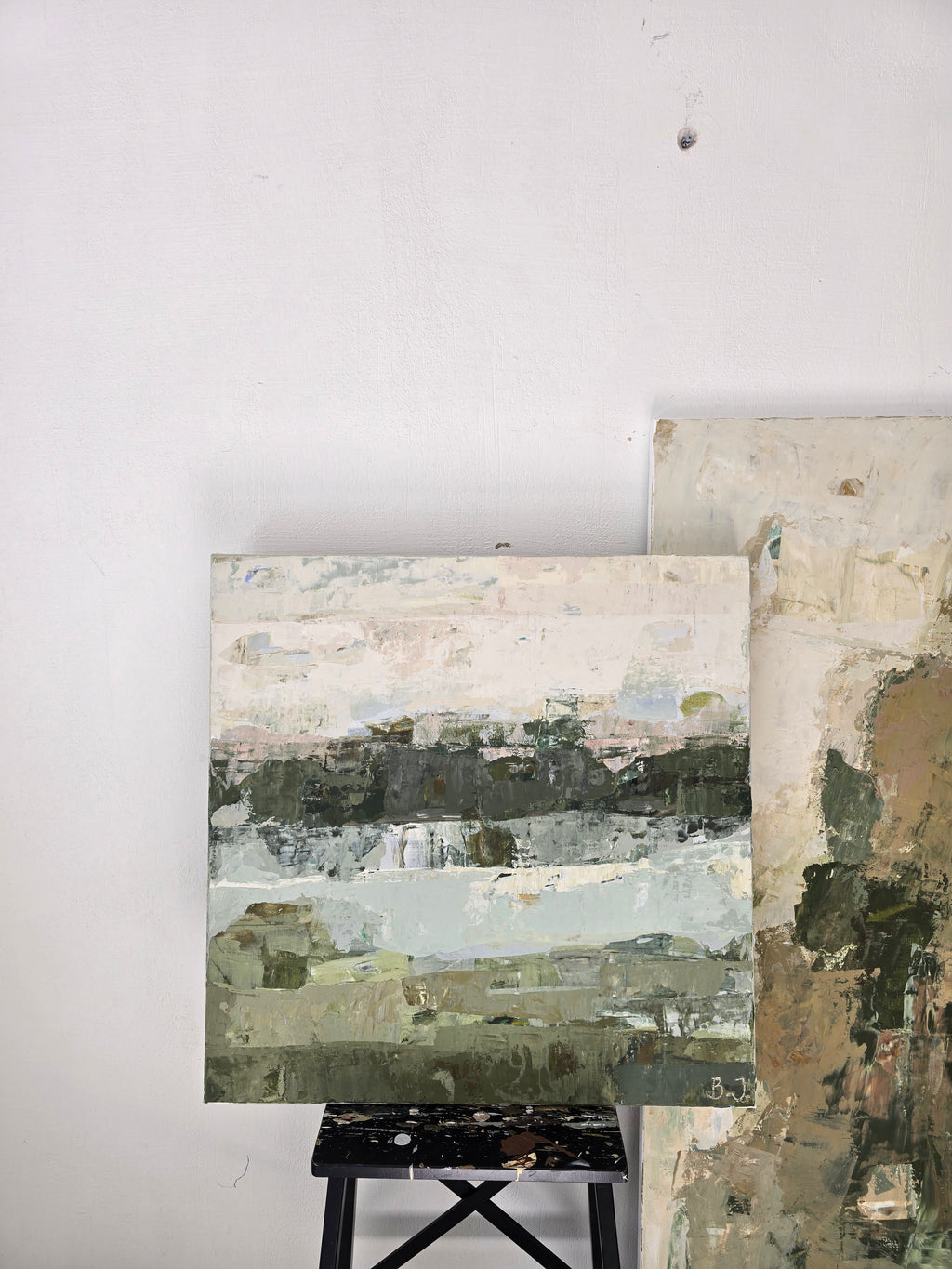 An abstract landscape painting created by Birute Studio on canvas displayed on a small black stand against a white wall, with another painting partially visible behind it.