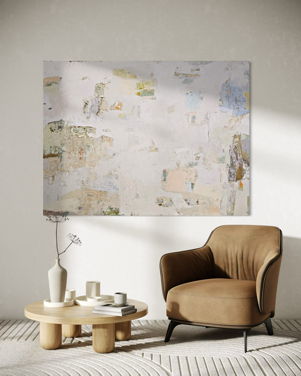 An abstract canvas painting with textured beige, cream, and light blue tones hangs on a light gray wall in a styled living room setting, featuring a brown armchair, a low wooden coffee table with ceramic vases, and a patterned rug.