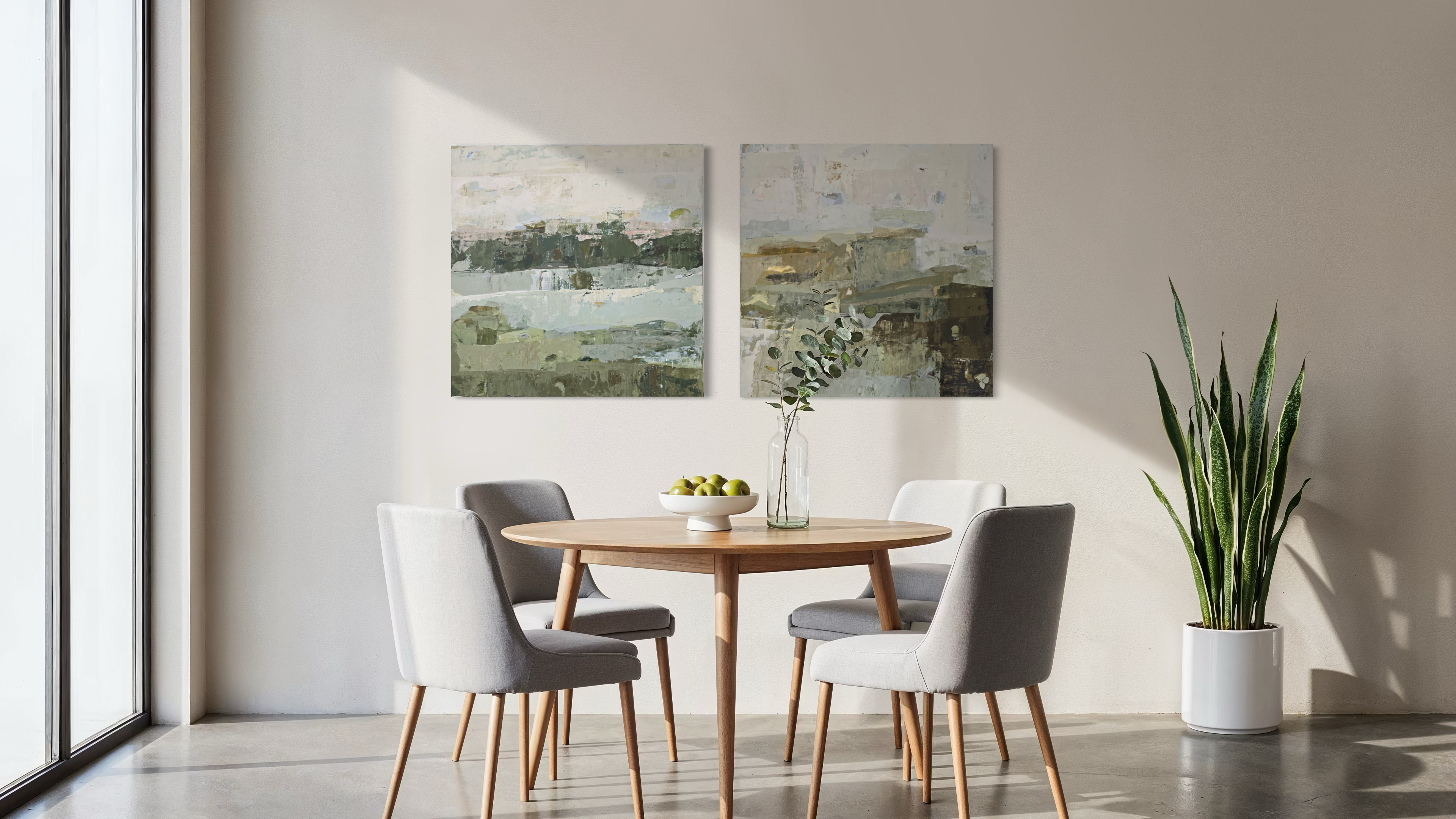 A styled dining room scene with two abstract landscape original paintings created by Birute Studio hanging above a round wooden table and four gray upholstered chairs. A potted snake plant stands on the floor to the right, and sunlight streams in from a large window on the left.