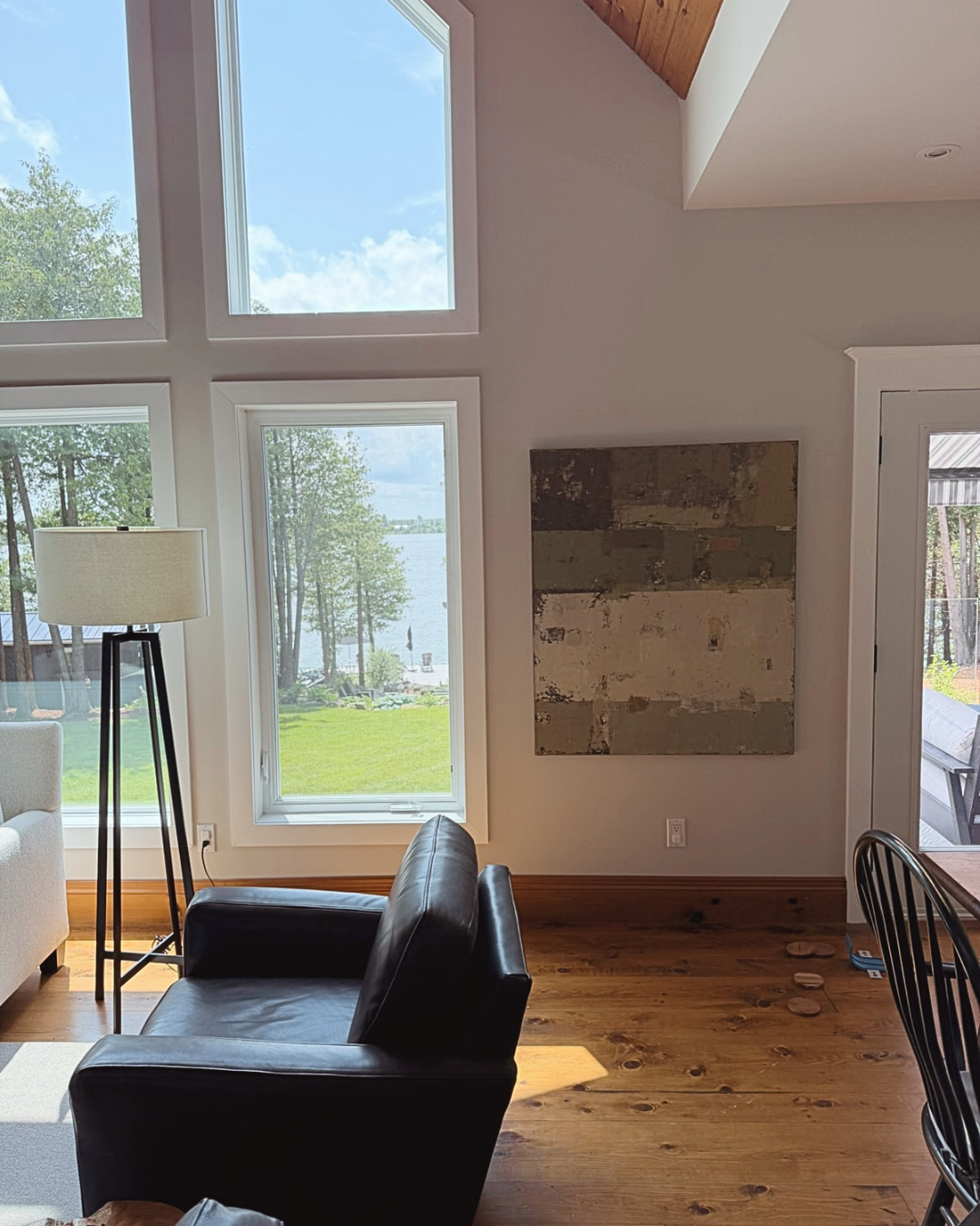 A modern living room interior with a black leather armchair in the foreground, a floor lamp, large windows overlooking a lake and trees, and an abstract wall art piece rich with layered texture created by Birute Studio.